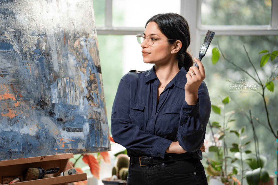 Contemplative artist with a paintbrush in hand standing in front of her abstract painting in a bright studio.