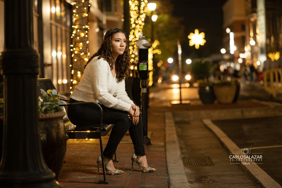 A young woman seated on a metal chair on a city sidewalk at night, with festive lights and street lamps illuminating the background.