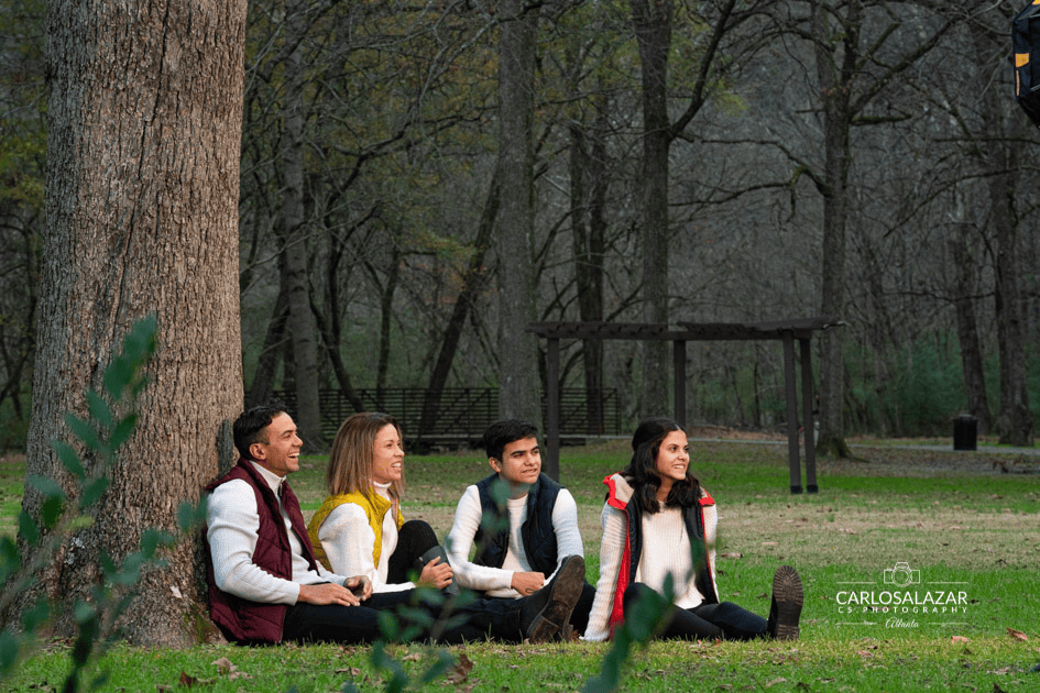 A family of four, including two adults and two teenagers, smiling and enjoying an autumn day in the park, seated on the grass next to a large tree.