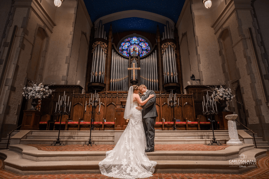 A bride and groom embracing at the altar in a church, with a large pipe organ and a stained glass window in the background.